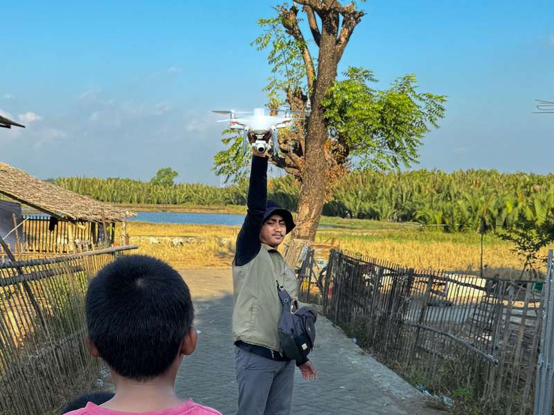Man holds a drone above his head while a child watches on a rural path near fields and water.