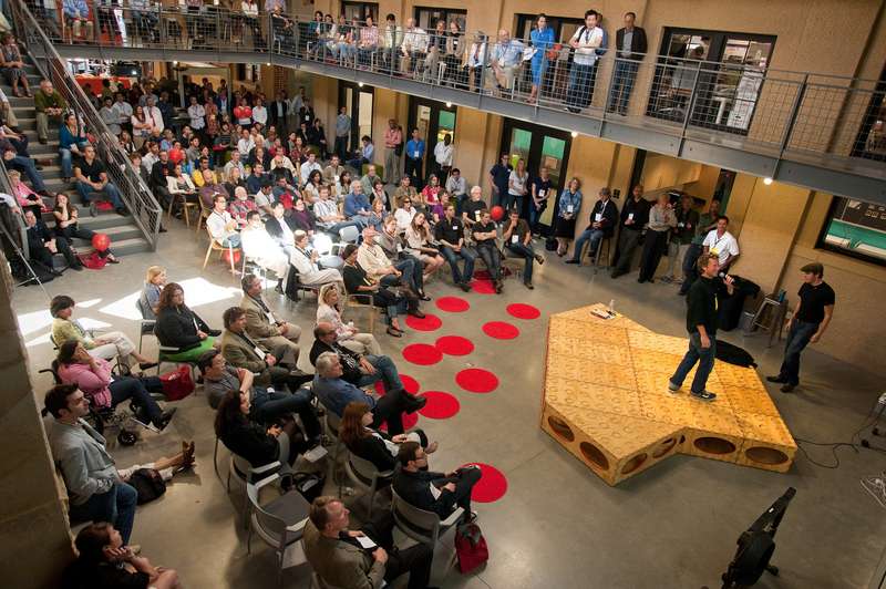 An audience gathers in the d.school for the opening ceremony, while a speaker stands on a large wooden stage, engaging attendees.