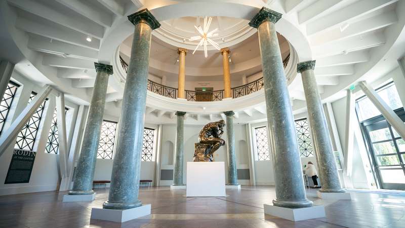 Rodin's "The Thinker" bronze sculpture on a white pedestal inside the Cantor Arts Center rotunda, surrounded by tall marble columns beneath a domed ceiling.