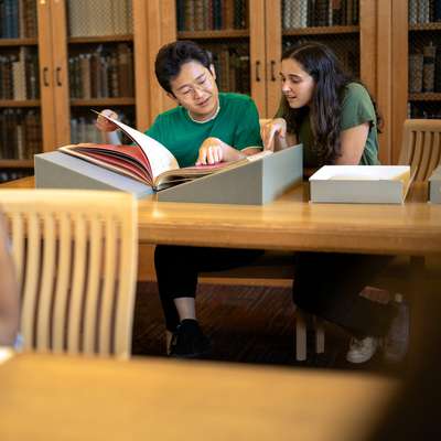 Students collaborate over a large book in a library while another student reads at a separate table.