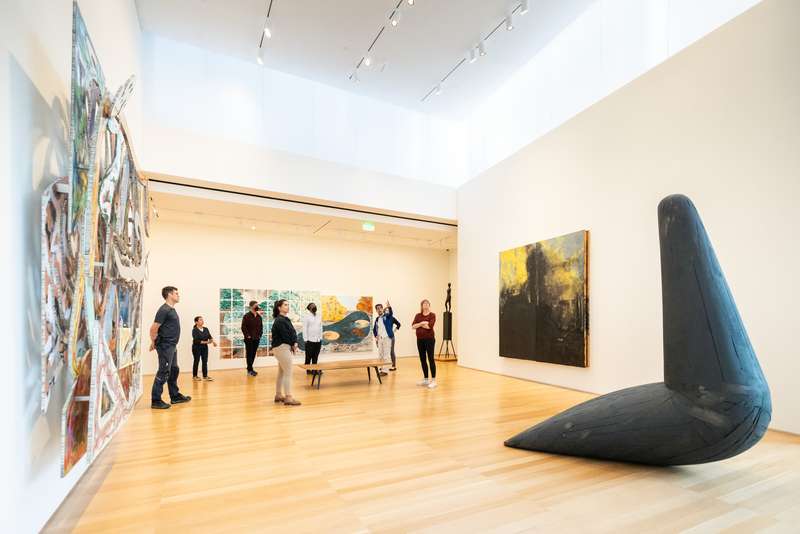 Visitors explore an art gallery featuring colorful paintings and a large black sculpture on the floor, enjoying the contemporary space.