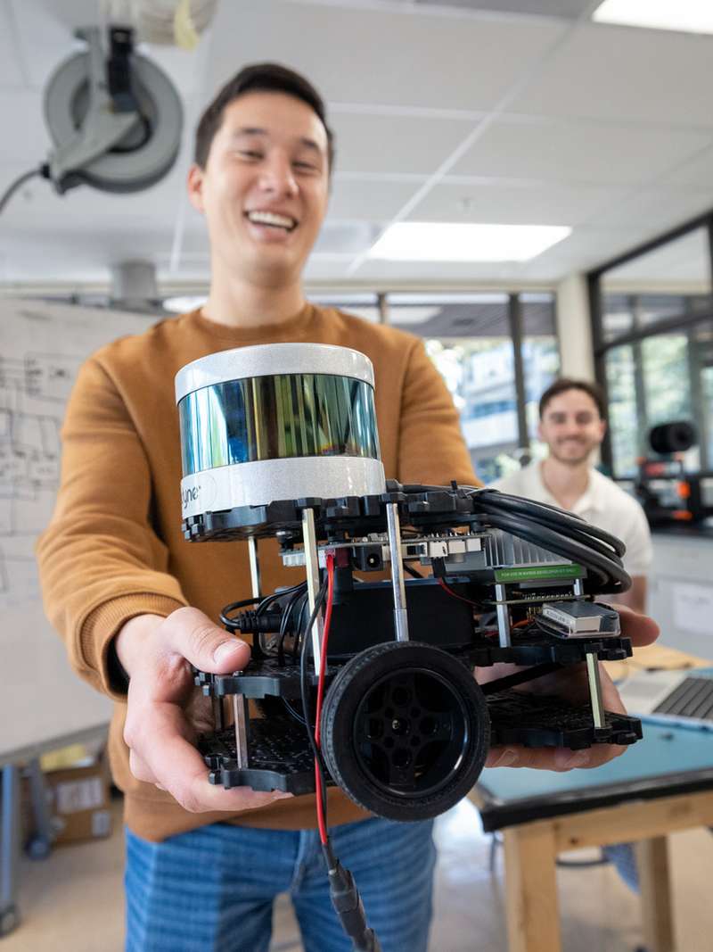 Grayson Armour holds a robotic device with sensors and electronics, while two peers observe in a lab setting.