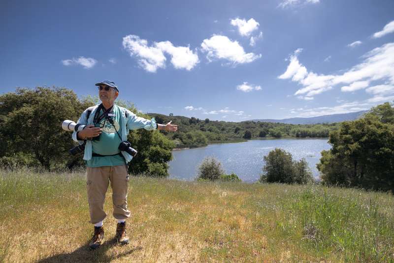 A Jasper Ridge docent gestures while giving a tour by a serene lake, surrounded by greenery on a sunny day.