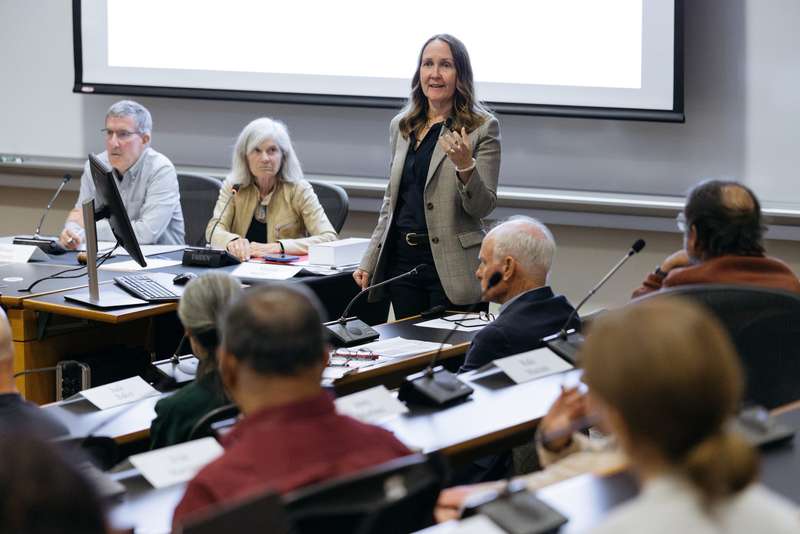Jenny Martinez presents to the Faculty Senate in a classroom, with panelists seated at the front discussing various topics.