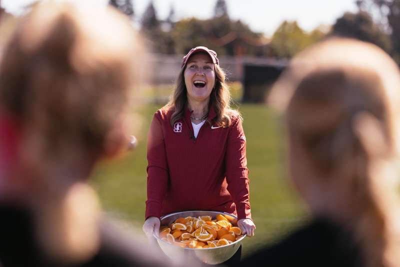 Jenny Martinez holds a bowl of oranges while speaking to the Stanford women's soccer team on a sunny field.