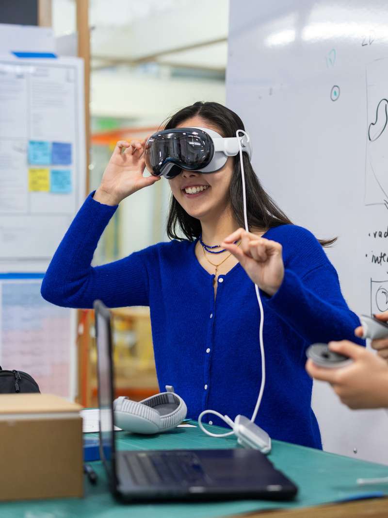 Two people interact with virtual reality equipment in a classroom, with a whiteboard filled with notes and diagrams in the background.