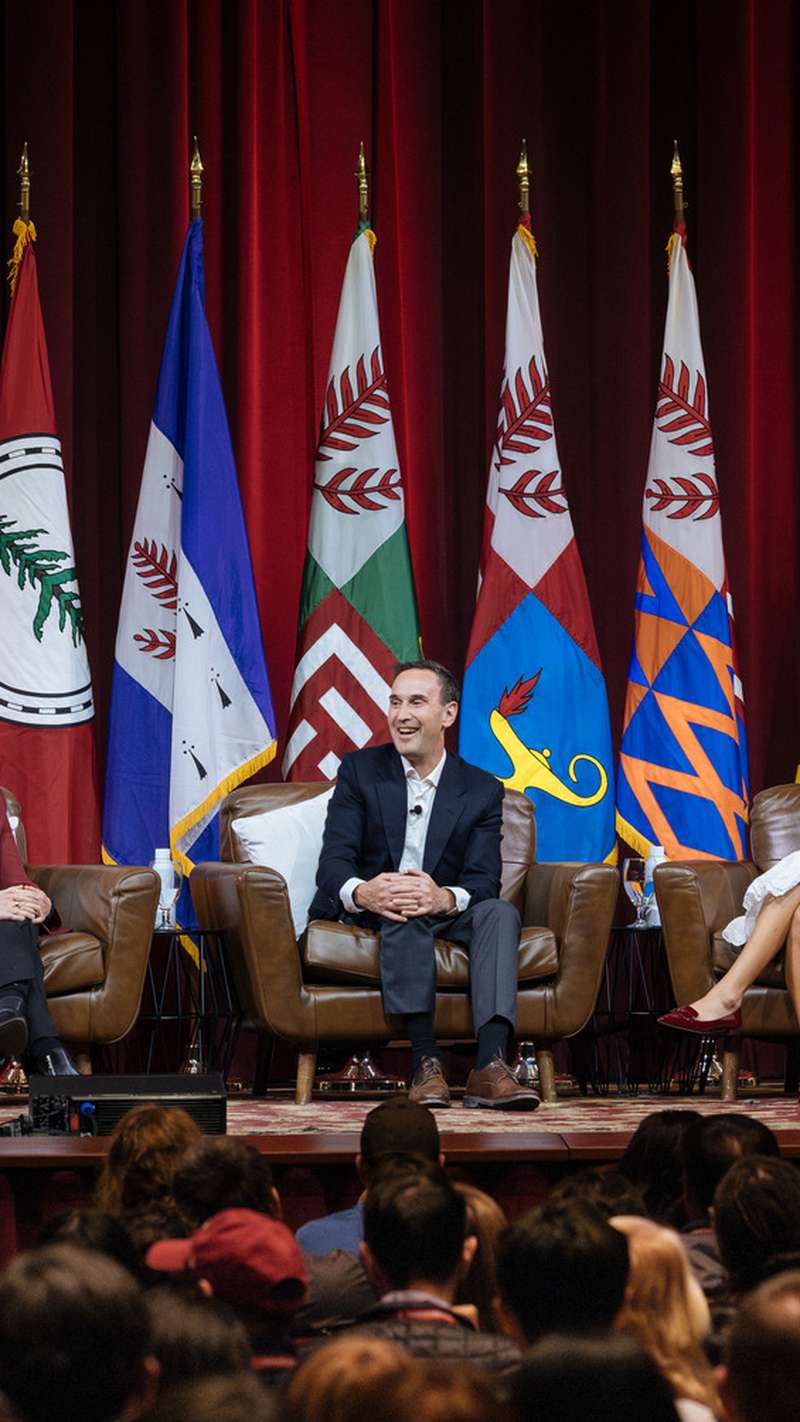 A panel discussion with four speakers seated on couches, facing an audience, with multiple flags displayed in the background.