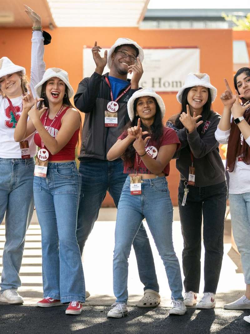 Seven cheering Stanford student orientation leaders in white fur hats and red shirts strike playful poses outside a dorm during new-student move-in, one waving a flag.