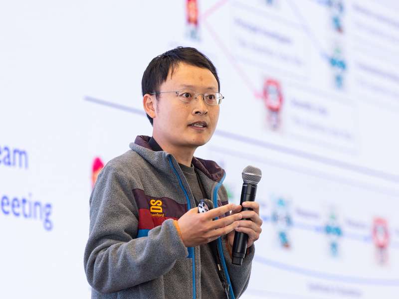 James Zou holding a microphone and gesturing while speaking at an event.