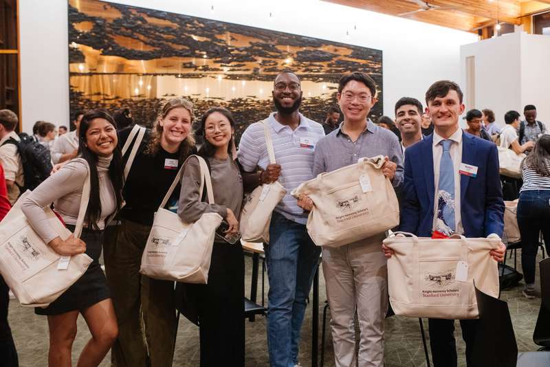 A group of six people poses with tote bags featuring a Stanford University Knight-Hennessy Scholars logo during a social event.