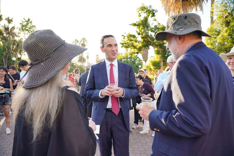 President Levin converses with two people at an outdoor event, with palm trees and a crowd in the background.