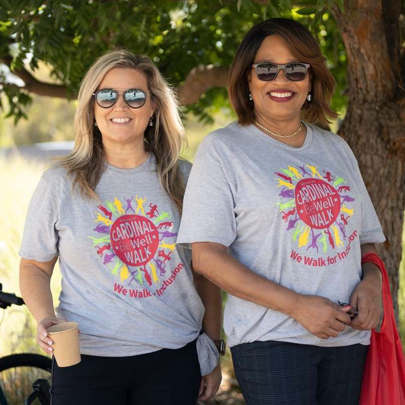 Two women smiling outdoors under a tree, wearing matching gray "Cardinal Be Well Walk – We Walk for Inclusion" t-shirts. A bicycle stands beside them.