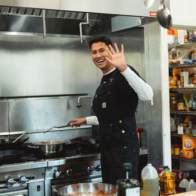 Adam Nayak stands in a busy kitchen, waving while cooking on the stovetop, with shelves of ingredients in the background.