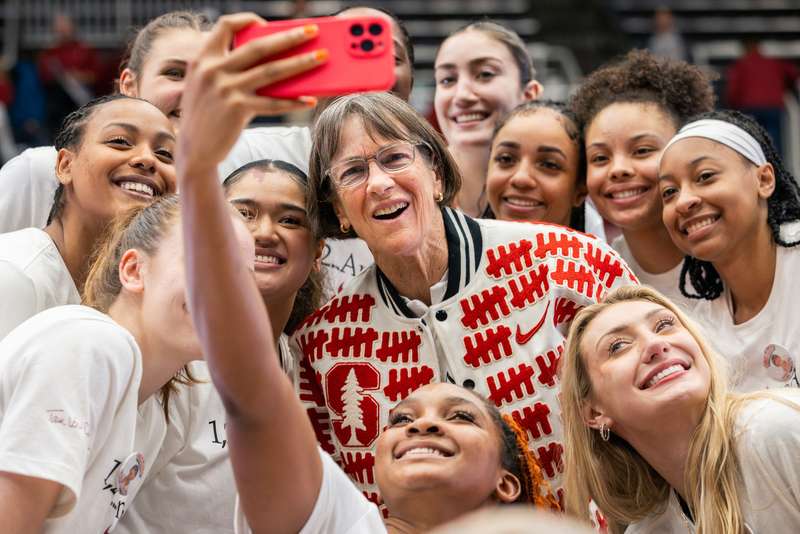 Coach Tara VanDerveer, wearing a jacket with tally marks on it, poses for a selfie with a group of Stanford women's basketball players
