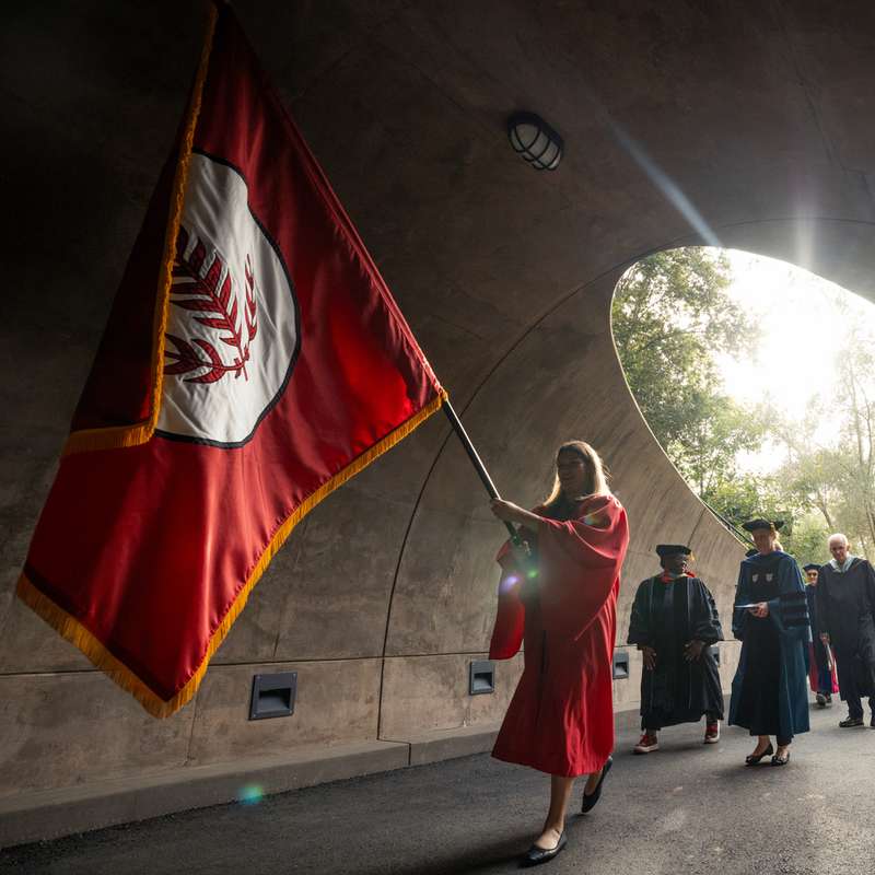 Students and faculty in ceremonial robes walk through an archway, carrying a large flag, during Convocation.
