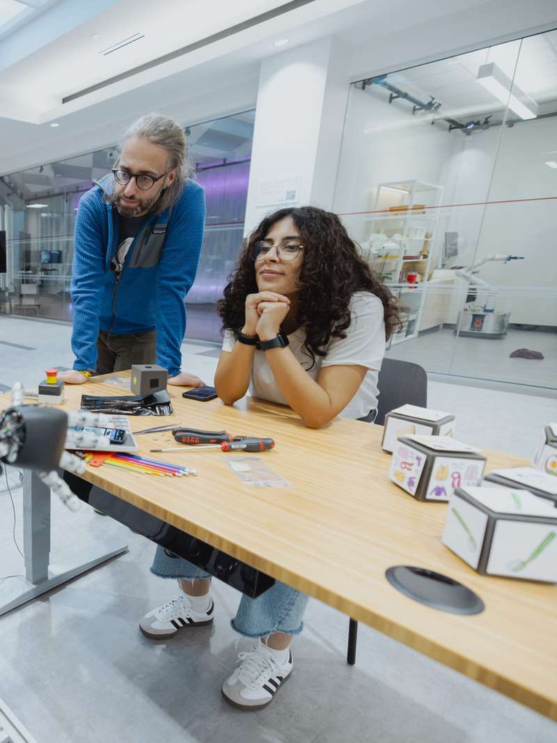 Three people work together on robotic components at a table in a modern lab, with another robot standing in the background.
