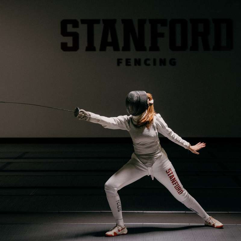 Liana Keesing in fencing gear and practicing with a sword in a dimly lit fencing gym.