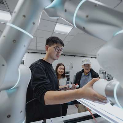 A person demonstrates a robotic arm's functionality to two observers in a modern lab setting.