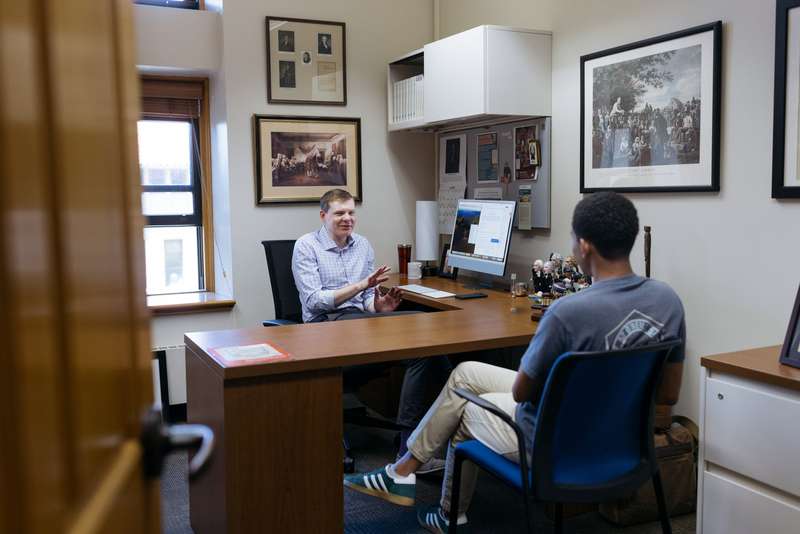 Professor Jonathan Gienapp and Diego Kagurabadza engage in a conversation at a desk in an office, surrounded by framed photos and a computer.
