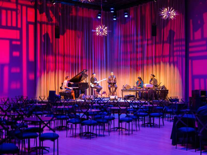 A small ensemble with piano, bass, and violin rehearses on a stage lit pink and purple, before empty cabaret-style seating.