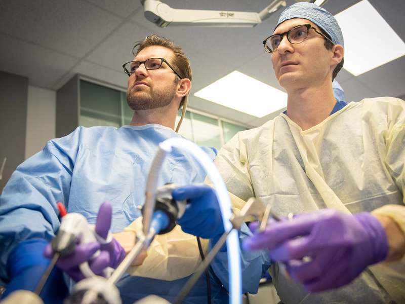 Two people in gowns and gloves look up toward a monitor while maneuvering surgical instruments, photographed from a low angle beneath the surgical tools.