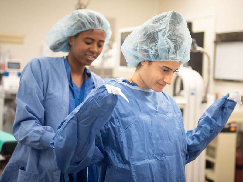 Two medical students in surgical attire assist each other in putting on protective gowns in a medical setting.