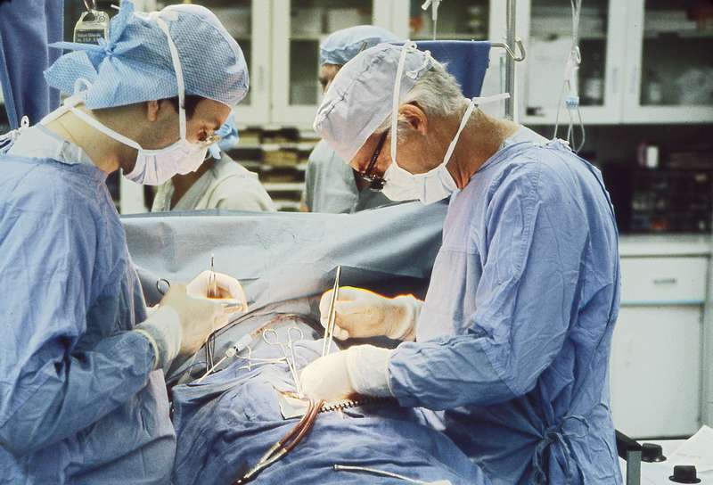 Dr. Norman Shumway and another doctor wearing scrubs and operating on a patent in a medical facility with other medical staff in the background.