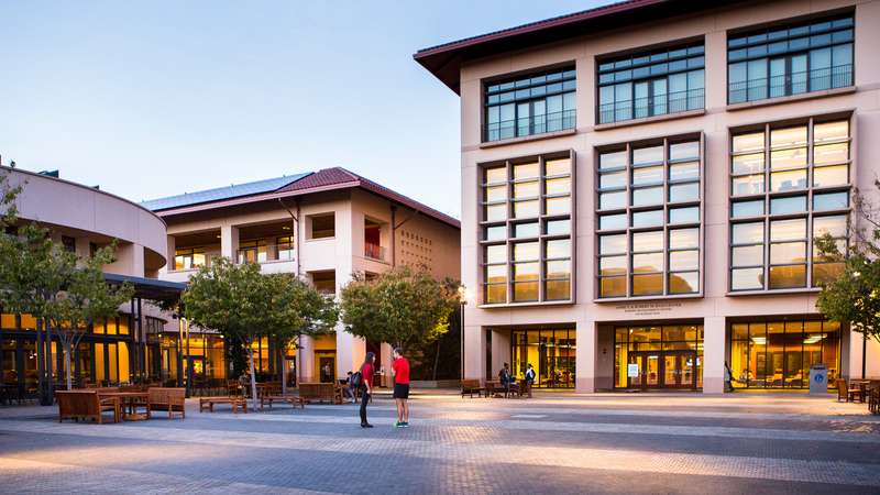 Two people talk in a university courtyard surrounded by modern buildings and trees, with students sitting on benches nearby.