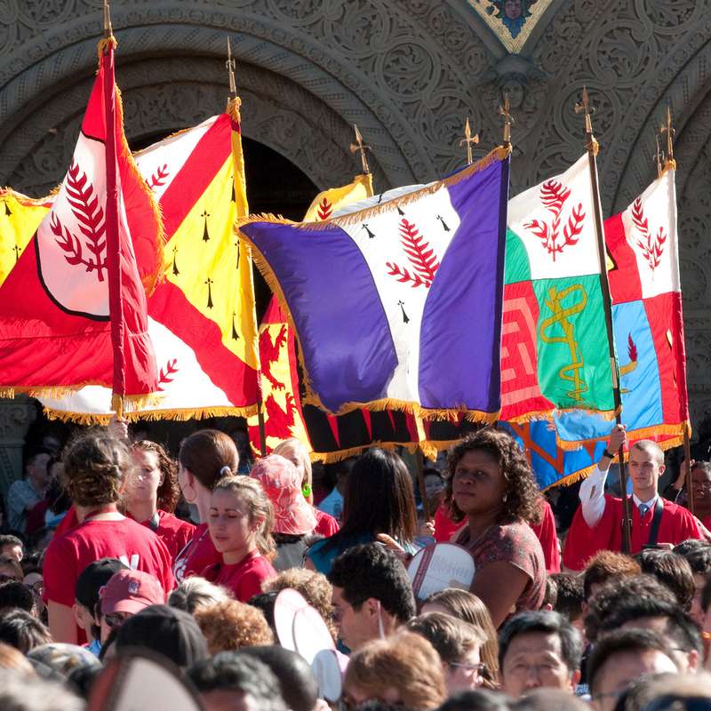 Graduates carry school banners during a commencement ceremony, surrounded by an audience celebrating the occasion.