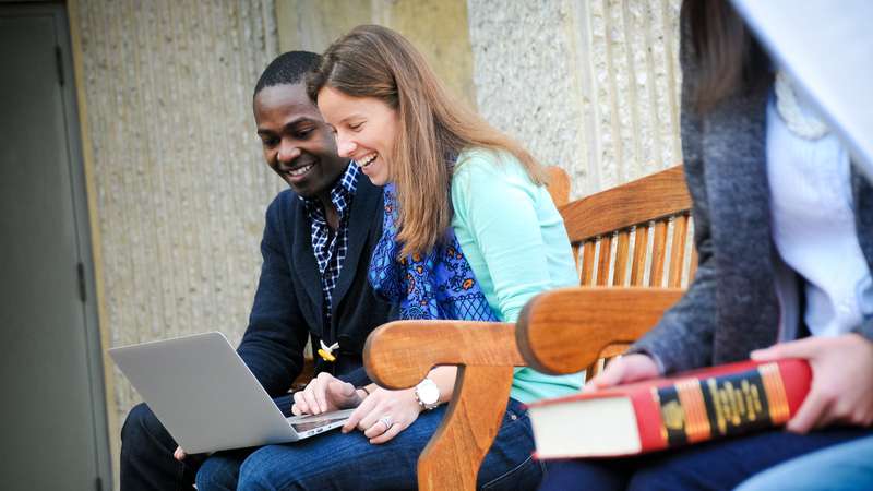 Two students collaborate on a laptop while seated on a bench, with another student reading a book nearby.