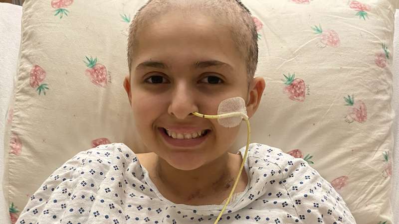 A young girl with shaved head, wearing a hospital gown and with a tube in her nose, smiles at the camera