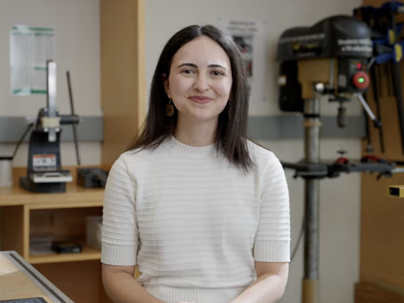 Aya Mouallem smiles at the camera in a workshop, with a drill press and clamps visible behind her.