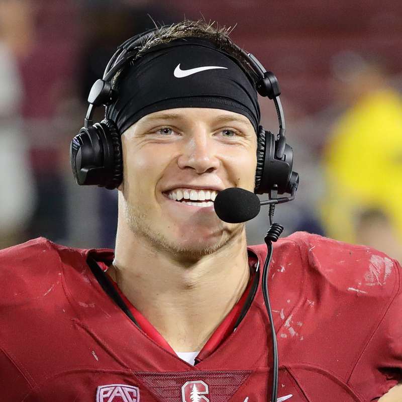 Christian McCaffrey in a Cardinal football jersey and pads, wears a headset and microphone and smilies while focused on the field.