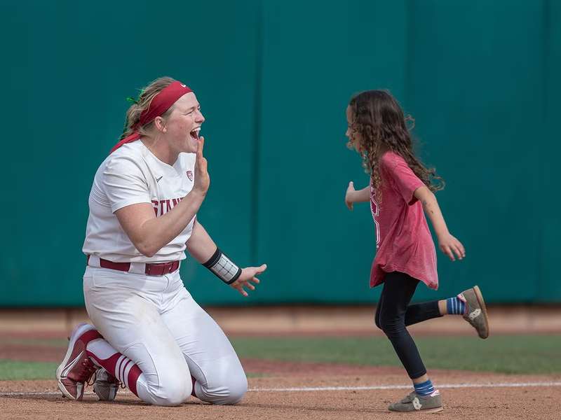 A softball player kneels with a smile and holds her hand out for a high-five as a young girl runs toward her on the field during a game.