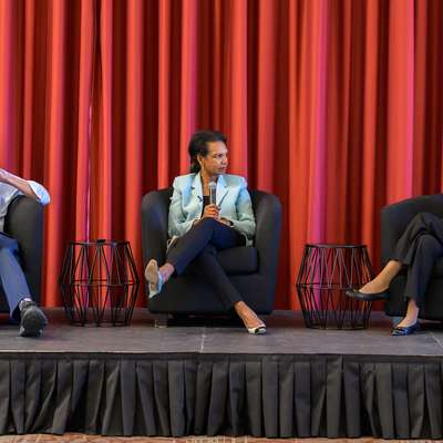 Three panelists seated in armchairs on a stage with a red curtain backdrop, holding microphones during a discussion.