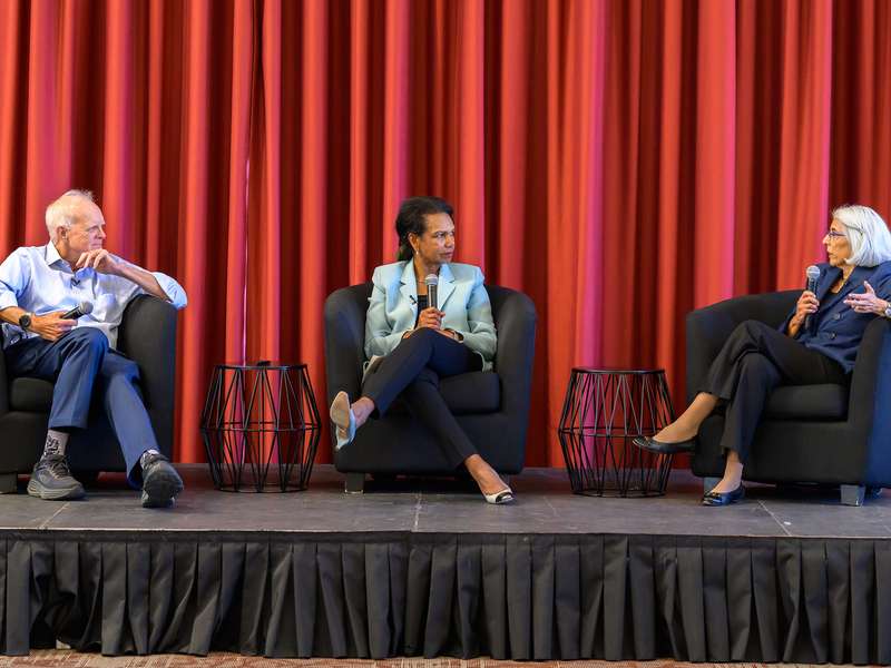 Three panelists seated in armchairs on a stage with a red curtain backdrop, holding microphones during a discussion.