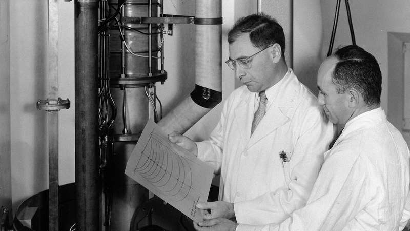 Two scientists in lab coats examine a paper with diagrams while standing beside a linear accelerator in a laboratory.
