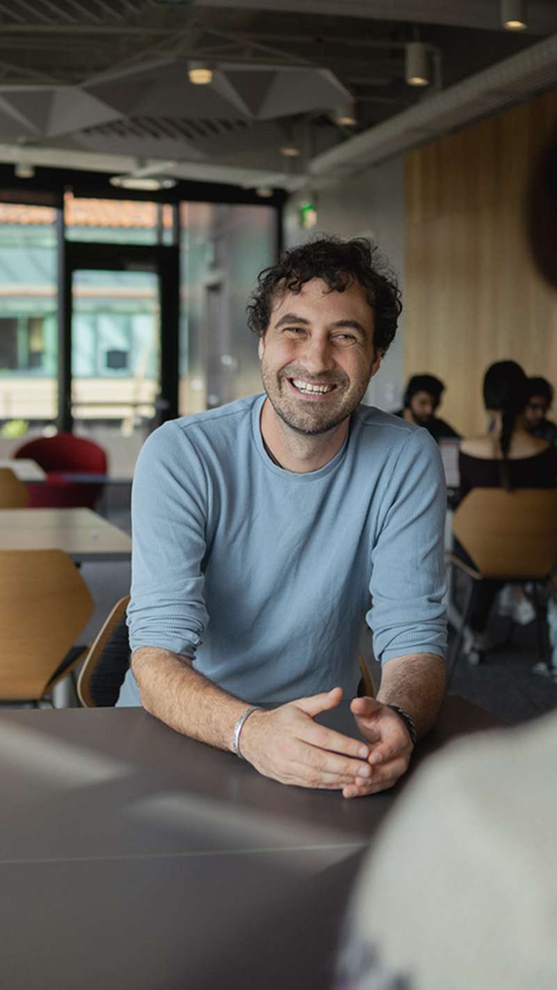 A group of four people engage in conversation around a table in a modern, well-lit meeting space, with two men prominently featured.