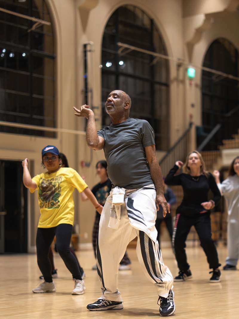 A dance class with multiple students practicing choreography in a spacious studio with bright lighting and wooden floors.