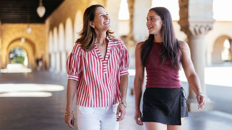 Two people engaged in conversation, walking together past sandstone columns on the Stanford campus.