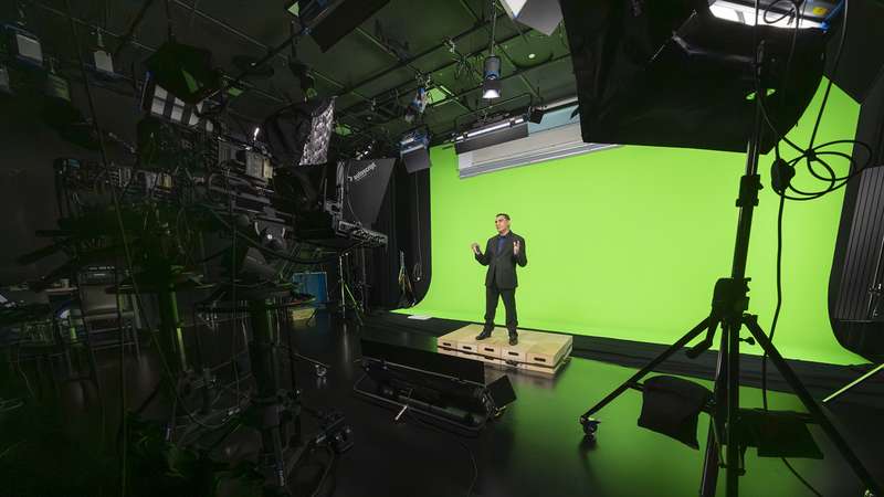 A presenter in a suit stands on a platform in front of a green screen setup, surrounded by studio lighting and equipment.