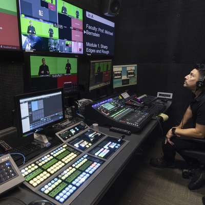 A person monitors multiple screens and a control panel in a studio, preparing for a live broadcast on green screen content.