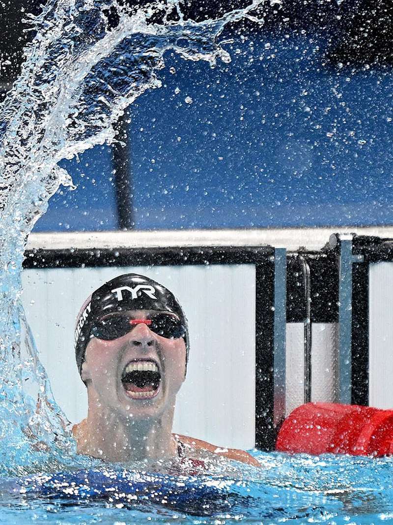 Swimmer Katie Ledecky shouts in celebration in the pool amid a big splash of water, wearing a cap and goggles, at the Paris 2024 Olympic Games.