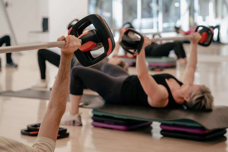 Group fitness class lying on mats, pressing weighted barbells upward in a body-pump-style strength workout in a bright studio.