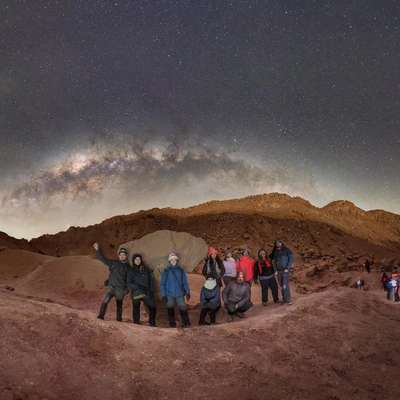 A group of people poses under a starry sky with the Milky Way visible above a desert landscape at night.