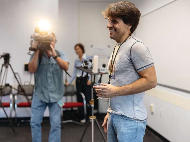 Karl Deisseroth holding a plastic champagne coupe stands near news microphones as a TV camera operator films.