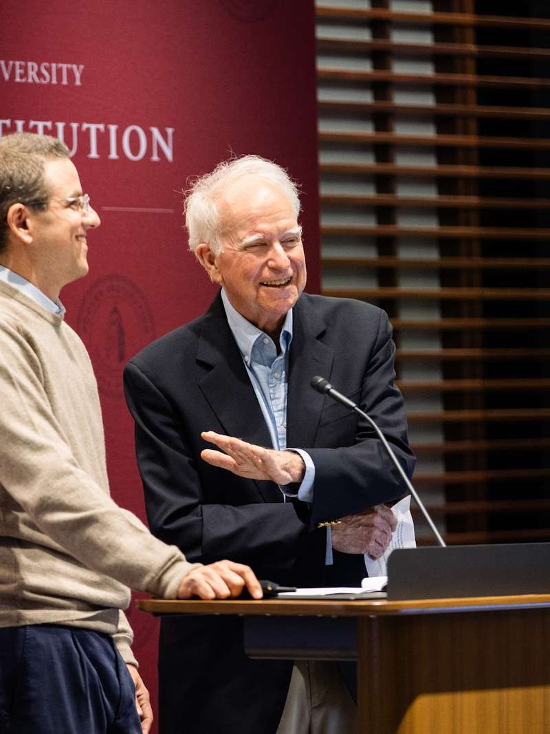 Robert E. Hall smiles at a podium in front of a backdrop that has the words "Stanford University" and "Hoover Institution" on it