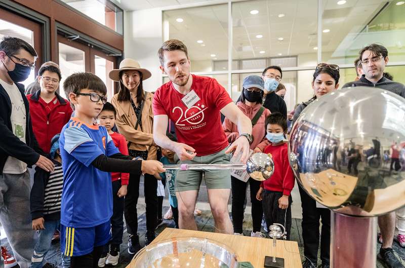 A presenter demonstrates an experiment to children and adults at a science fair, engaging them with hands-on learning activities.