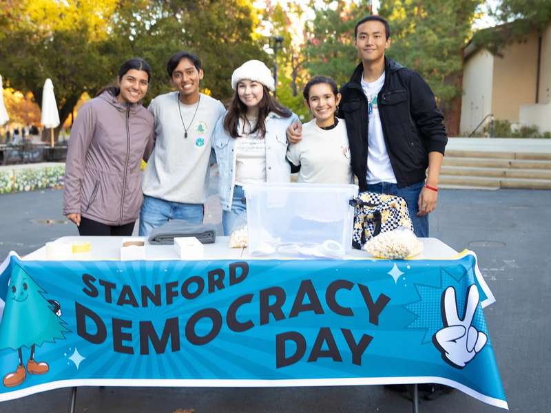 Five students smile together behind a table draped with a Stanford Democracy Day banner on an outdoor campus plaza.
