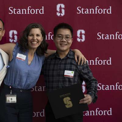 Three individuals celebrate with arms around each other, holding certificates in front of a Stanford University backdrop.